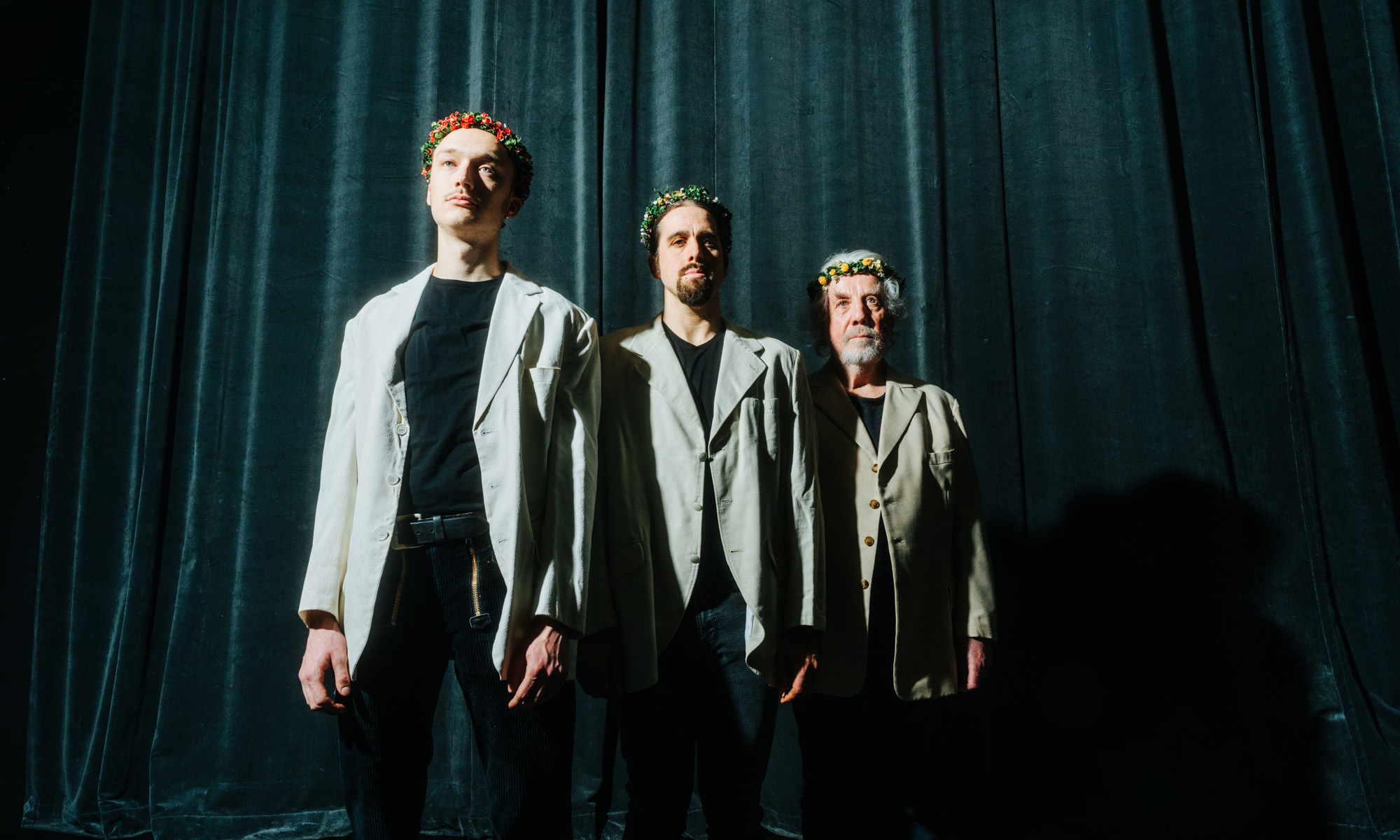 Drei Männer unterschiedlichen Alters stehen vor einem dunkelblauen Vorhang und tragen Blumenkränze auf ihren Köpfen.